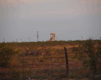 [An animated sequence of two images of a yellow oil derrick as it pumps in a field beyond a barbed-wire fence.]